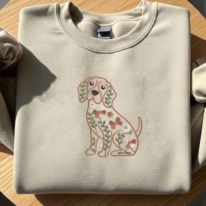 May include: Beige sweatshirt featuring an embroidered dog design with floral accents in shades of brown, green, and white. The dog is sitting, with the floral design covering its body. The sweatshirt has a crew neck and long sleeves.