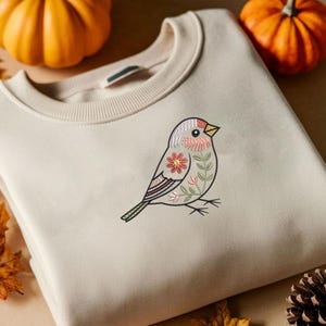 Puede incluir: Sudadera beige con un dise&ntilde;o de p&aacute;jaro bordado. El p&aacute;jaro tiene la cabeza gris, detalles rosas y un estampado floral en el cuerpo. El dise&ntilde;o incluye una flor roja y hojas verdes. La sudadera est&aacute; doblada, con calabazas en el fondo.