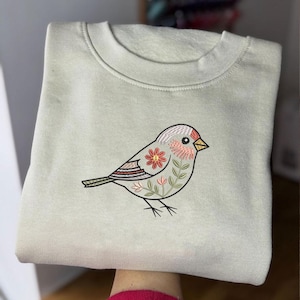 Puede incluir: Una sudadera doblada, de color verde claro, con un dise&ntilde;o de p&aacute;jaro bordado. El p&aacute;jaro tiene una cabeza roja, rosa y blanca, una flor roja en el cuerpo y hojas verdes. La sudadera est&aacute; hecha de un material suave.