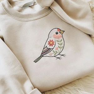 Puede incluir: Una sudadera de color crema con un dise&ntilde;o de p&aacute;jaro bordado. El p&aacute;jaro est&aacute; adornado con patrones florales y de hojas en tonos de rojo, rosa y verde. El p&aacute;jaro tiene un pico amarillo y est&aacute; posado sobre finas l&iacute;neas negras.