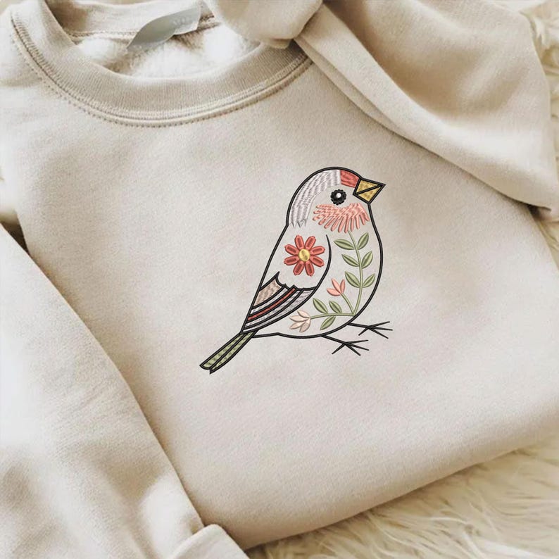 Puede incluir: Una sudadera de color crema con un dise&ntilde;o de p&aacute;jaro bordado. El p&aacute;jaro est&aacute; adornado con patrones florales y de hojas en tonos de rojo, rosa y verde. El p&aacute;jaro tiene un pico amarillo y est&aacute; posado sobre finas l&iacute;neas negras.