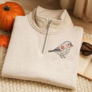 Puede incluir: Sudadera beige claro con cremallera de cuarto de cuello y dise&ntilde;o de p&aacute;jaro bordado con flores rojas y rosas y hojas verdes. La sudadera tiene cuello alto y cremallera plateada. Una peque&ntilde;a calabaza naranja en el fondo.