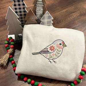 Puede incluir: Sudadera crema con un dise&ntilde;o de p&aacute;jaro bordado. El p&aacute;jaro presenta un patr&oacute;n floral con acentos rojos, rosas y verdes. &Aacute;rboles de Navidad decorativos de madera y una guirnalda de cuentas en el fondo.