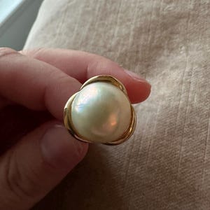 May include: A gold-toned ring featuring a large, iridescent pearl. The pearl is round and has a creamy white color with subtle hints of pink and green. The ring's setting has a three-petal design.