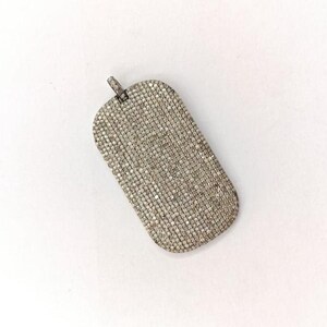 May include: A rectangular pendant with a loop at the top, covered in small, sparkling stones. The pendant has rounded edges and a textured surface, reflecting light. The overall color is a light silver or gray.