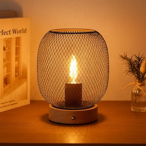 Peut inclure: Une lampe de table décorative avec un abat-jour en treillis métallique noir et une base en bois. La lampe émet une lumière chaude et vacillante, créant une ambiance chaleureuse. La lampe est sur une surface en bois.