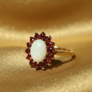 May include: A gold ring with an oval, white opal centre stone, encircled by small, round, red gemstones. The ring's band is plain gold. The ring is set against a gold background.