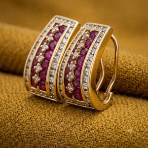 May include: A pair of gold hoop earrings featuring a central row of round, deep red rubies flanked by two rows of small, clear white diamonds. The earrings have a curved, rectangular shape with a polished gold finish and a secure hinged backing.