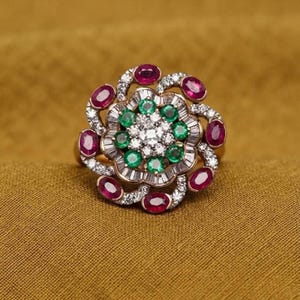 Peut inclure: Une bague en forme de fleur avec un groupe central de petits diamants blancs ronds entouré d'un cercle d'émeraudes vertes rondes. Le bord extérieur est orné de diamants taille baguette et de rubis rouges ovales sur une monture métallique.