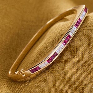 Puede incluir: Un brazalete de oro con un diseño rectangular. El brazalete está adornado con filas alternas de piedras preciosas rojas y blancas. El brazalete descansa sobre una tela dorada.