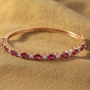 May include: A rose gold bangle bracelet featuring an alternating pattern of oval-cut red rubies and round-cut clear white diamonds set in a continuous circular band with a secure hinged clasp mechanism.