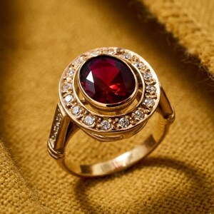 Puede incluir: Un anillo de oro con una gran piedra preciosa roja oscura de corte ovalado en el centro, rodeada por un halo de piedras preciosas más pequeñas y claras. La banda y el engaste del anillo también son de oro, creando una apariencia lujosa y elegante.