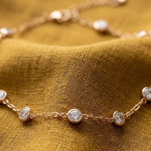 May include: A delicate gold bracelet with multiple clear, round gemstones. The bracelet is set against a textured, golden-yellow background, highlighting the sparkle of the gems and the warm tone of the metal.