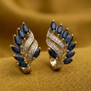 May include: A pair of gold-toned earrings featuring a leaf-like design. Each earring has a series of dark blue, oval-shaped stones and a central band of smaller, clear stones. The earrings are set against a golden-yellow background.