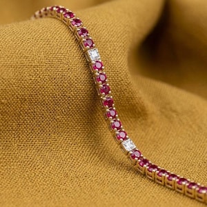 May include: A gold tennis bracelet with alternating round ruby gemstones and square-cut diamond accents. The bracelet is set against a golden-brown fabric background, highlighting the jewelry's sparkle and color.