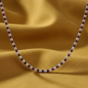 May include: A delicate necklace featuring alternating red and clear gemstones. The gemstones are set in a silver or white metal chain. The necklace is displayed against a golden-yellow, textured background.