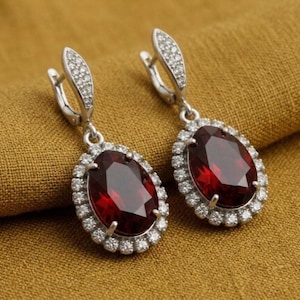 Può includere: Un paio di orecchini pendenti color argento. Ogni orecchino presenta una grande gemma rosso scuro, ovale e sfaccettata, circondata da un alone di piccole gemme chiare. La parte superiore di ogni orecchino ha un design curvo con altre piccole gemme chiare.