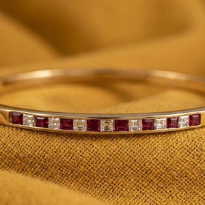 Pode incluir: Uma pulseira de ouro com pedras preciosas vermelhas e brancas quadradas alternadas. A pulseira é exibida em um fundo de tecido amarelo dourado, destacando o design e a cor da joia.