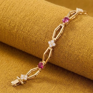 May include: A gold-toned bracelet featuring a series of elongated, open geometric links. The design alternates between small, square-shaped clusters of clear white stones and single, round, vibrant pink stones set within the metal framework.