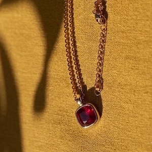 May include: A gold-toned necklace with a square, deep red gemstone pendant. The pendant is set in a silver-colored metal. The chain is delicate and hangs against a mustard-colored background.