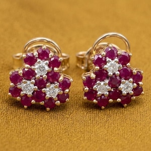 May include: A pair of gold-toned stud earrings. Each earring features a floral design with deep red gemstones and sparkling white stones. The earrings are set against a golden-brown background.