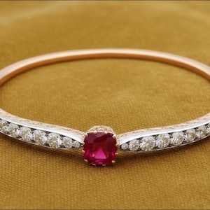 May include: A rose gold and silver bracelet with a large, square, ruby-colored gemstone at the center. The bracelet features a row of small, clear gemstones on each side of the ruby. The background is a solid, golden-brown color.
