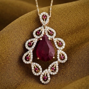 May include: A gold pendant necklace featuring a large pear-shaped deep red ruby at the centre, surrounded by a decorative frame of smaller round rubies and numerous tiny white diamonds set in a filigree-style metal arrangement on a gold chain.