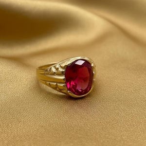 May include: A gold ring with a large, oval-shaped, ruby-red gemstone. The ring has a detailed, textured design on the band and around the gemstone setting. The ring is displayed on a gold-colored fabric.