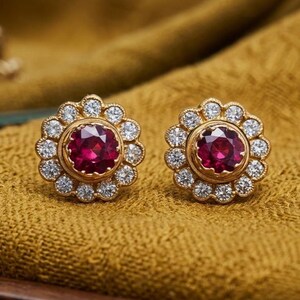 Puede incluir: Un par de pendientes de oro con diseño floral. Cada pendiente presenta una gema grande, redonda y de color rojo intenso, rodeada por un halo de pequeñas gemas transparentes. Los pendientes están colocados sobre una tela de color amarillo dorado.