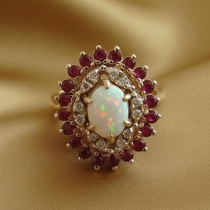 May include: A gold ring featuring an oval opal center stone surrounded by a halo of small red gemstones and clear accent stones. The ring's design is set against a gold background, highlighting the ring's intricate details and vibrant colors.