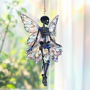 May include: A hanging ornament featuring a skeleton fairy with iridescent wings and a skirt. The fairy is black with a detailed skeletal structure. The wings and skirt have a stained glass effect. The ornament hangs from a silver chain.