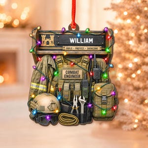 May include: A decorative ornament featuring a combat engineer vest, helmet, and tools. The top sign displays the name WILLIAM and the text BUILD PROTECT OVERCOME. The gear is wrapped in colorful string lights, hanging from a red ribbon.