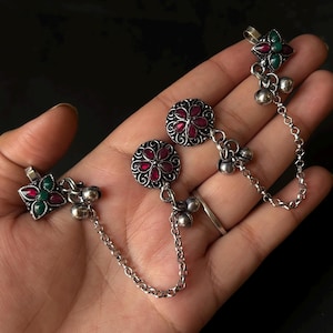 Op de afbeelding: Zilverkleurige oorbellen met rode en groene edelsteenaccenten. De oorbellen hebben een bloemmotief met een centrale rode steen, omringd door kleinere rode stenen. Een ketting verbindt de oorbellen met een kleiner, bijpassend edelsteenstuk.