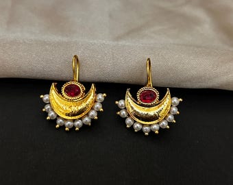 Pode incluir: Um par de brincos dourados. Cada brinco apresenta uma forma de meia-lua com uma pedra preciosa vermelha no centro e uma borda de pequenas pérolas brancas. Os brincos têm um fecho de gancho.