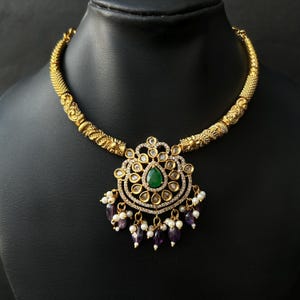May include: A gold-toned necklace with a central pendant featuring a green gemstone surrounded by clear stones. The pendant is adorned with small white and purple beads. The necklace is displayed on a black mannequin.