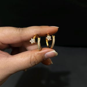 May include: A pair of gold-colored hoop earrings. Each earring features a small, sparkling flower design and is encrusted with small, clear gemstones. The earrings are held against a dark background.