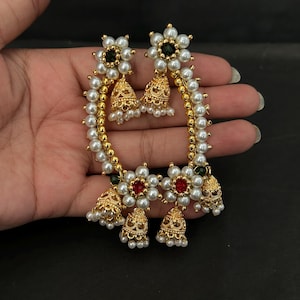 Puede incluir: Un par de pendientes dorados con un diseño curvo adornado con perlas artificiales blancas. Cada pendiente incluye dos motivos en forma de flor con piedras centrales rojas o verdes, acentuados por colgantes en forma de campana con cuentas.
