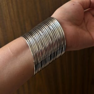 Può includere: Una collezione di braccialetti d'argento impilati su un polso. I braccialetti sono sottili, rotondi e lucenti, che riflettono la luce. I gioielli creano un look audace e stratificato. Lo sfondo è una superficie di legno sfocata.