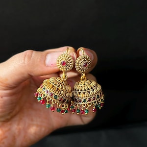 Op de afbeelding: Een paar goudkleurige jhumka-oorbellen. Elke oorbel heeft een koepelvormige belvorm met ingewikkelde filigraan details, versierd met rode en groene kralen. De oorbellen hebben een kleine, cirkelvormige bovenkant met een rood stenen midden.