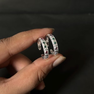 May include: A pair of silver hoop earrings featuring an intricate engraved pattern along the outer edge. Each hoop is accented with small, round, alternating green and red gemstones set into the metal, creating a colourful and detailed design.