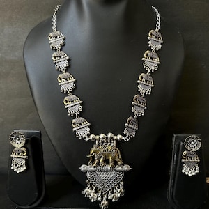 K&ouml;nnte beinhalten: Silberfarbenes Halsketten- und Ohrring-Set mit Elefantenmotiven. Die Halskette hat eine Kette mit dekorativen Elefantenanh&auml;ngern und einen gr&ouml;&szlig;eren Anh&auml;nger mit einem Elefanten-Design. Die Ohrringe spiegeln das Elefanten-Design wider.