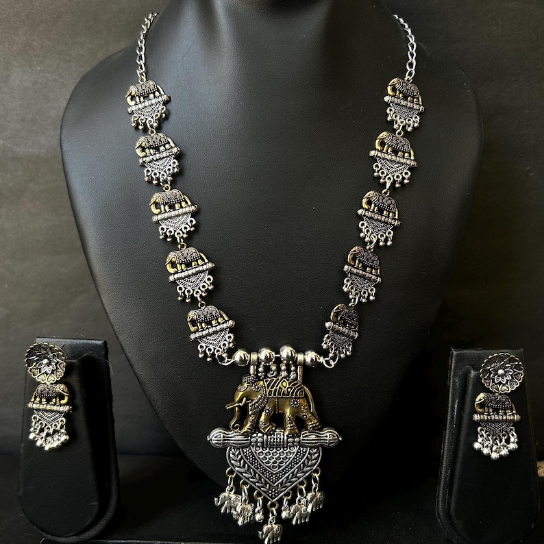 K&ouml;nnte beinhalten: Silberfarbenes Halsketten- und Ohrring-Set mit Elefantenmotiven. Die Halskette hat eine Kette mit dekorativen Elefantenanh&auml;ngern und einen gr&ouml;&szlig;eren Anh&auml;nger mit einem Elefanten-Design. Die Ohrringe spiegeln das Elefanten-Design wider.