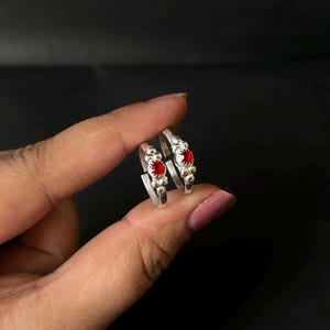 May include: A pair of silver toe rings, each with a central, bright red gemstone. The rings have a detailed design with small, beaded embellishments. The gemstones are round and set in a polished silver band.