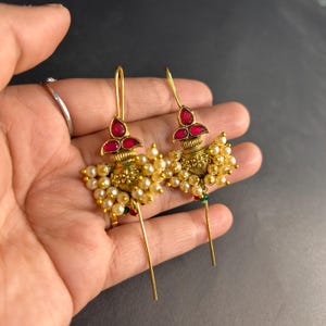 Op de afbeelding: Een paar goudkleurige oorbellen met rode edelsteenaccenten en clusters van kleine, crèmekleurige parels. De oorbellen hebben een gedetailleerd, sierlijk ontwerp met een lange, slanke haak. De oorbellen worden in een hand gehouden.