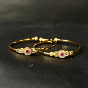 May include: Two gold bangles with a central design featuring a red gemstone surrounded by small white stones. The bangles have an ornate, floral-inspired pattern. They are set against a dark background.