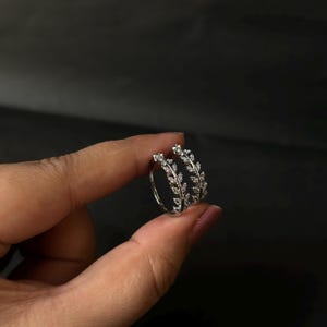 Puede incluir: Un par de pendientes de aro plateados. Cada pendiente presenta un delicado diseño de hojas adornado con pequeños cristales brillantes. Los pendientes se muestran sobre un fondo oscuro, resaltando sus intrincados detalles.