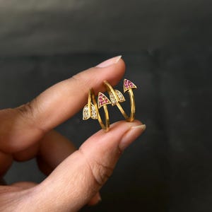 Op de afbeelding: Vier goudkleurige ringen met pijlvormige ontwerpen. Twee ringen hebben heldere stenen en twee hebben rode stenen. De ringen worden tegen een donkere achtergrond gepresenteerd.