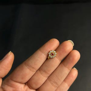 May include: A gold nose stud with a floral design. The centre of the flower is a red gemstone, encircled by clear, sparkling stones. The stud is small and delicate, suitable for everyday wear.