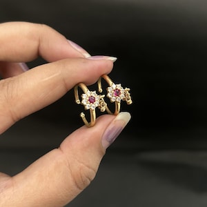 Puede incluir: Un par de anillos para el dedo gordo del pie dorados, cada uno con un diseño floral con una gema roja central rodeada de cristales transparentes. Los anillos se sostienen sobre un fondo oscuro, mostrando sus detalles.
