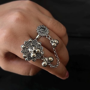 Könnte beinhalten: Ein silberfarbener Ring mit floralem Design und aufwendigen Details. Der Ring hat eine Kette, die mit einem kleineren Blumenelement am angrenzenden Finger verbunden ist, verziert mit kleinen, hängenden Silberperlen. Der Ring wird an einer Hand vor dunklem Hintergrund getragen.